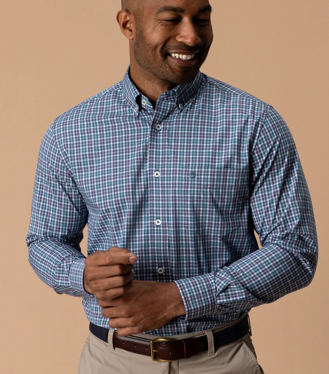 A man wearing a blue and purple plaid button-down shirt, paired with khaki pants and a brown leather belt, standing against a solid beige background.