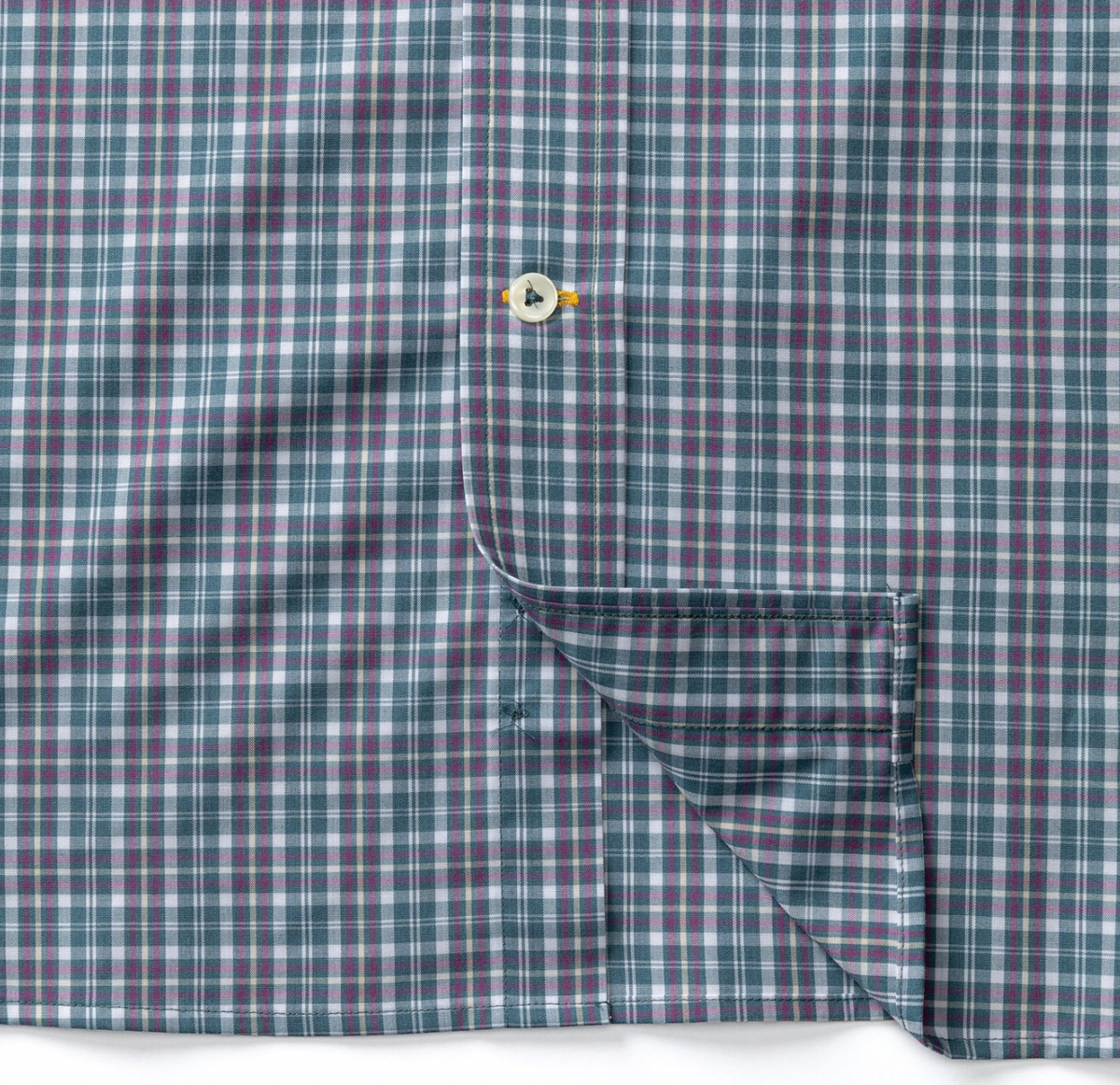 Close-up of a plaid button-up shirt fabric in muted teal, purple, and white, with a single button visible and a folded corner showing the pocket seam.