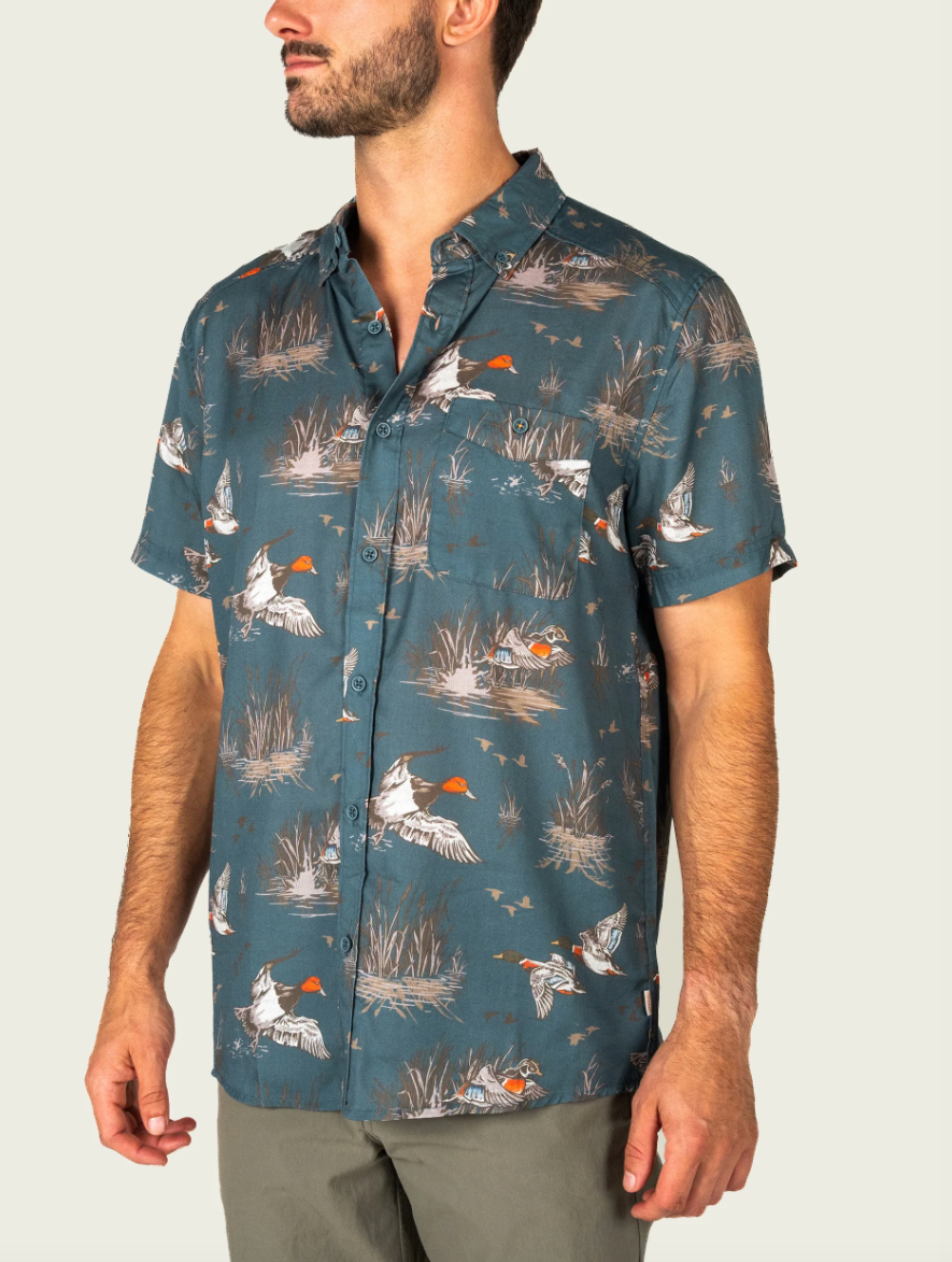 A man wearing a short-sleeve button-up shirt with a teal blue-green background and an allover print of ducks, reeds, and birds in white, orange, and gray, paired with gray pants against a light gray studio background.