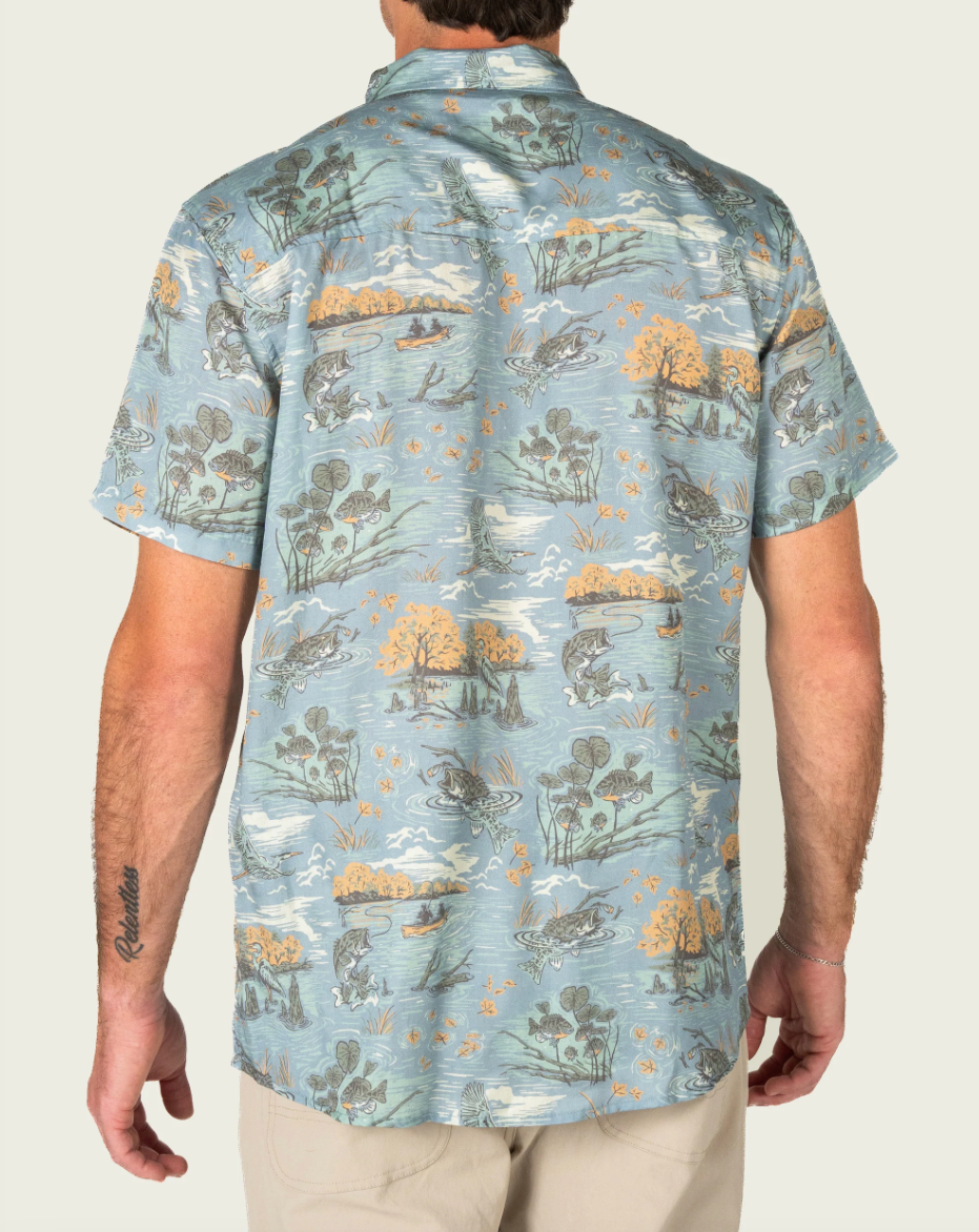 A man viewed from the back wearing a short-sleeve button-up shirt with a light blue, gray, and ochre landscape print featuring fish, trees, and reeds. He is also wearing khaki pants against a clean, off-white studio background.