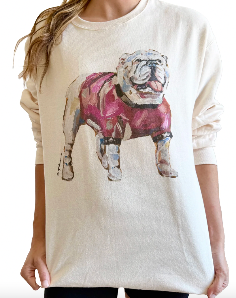 A woman wearing a cream-colored crewneck sweatshirt with a large acrylic-style graphic of an English Bulldog wearing a pink sweater on the front.
