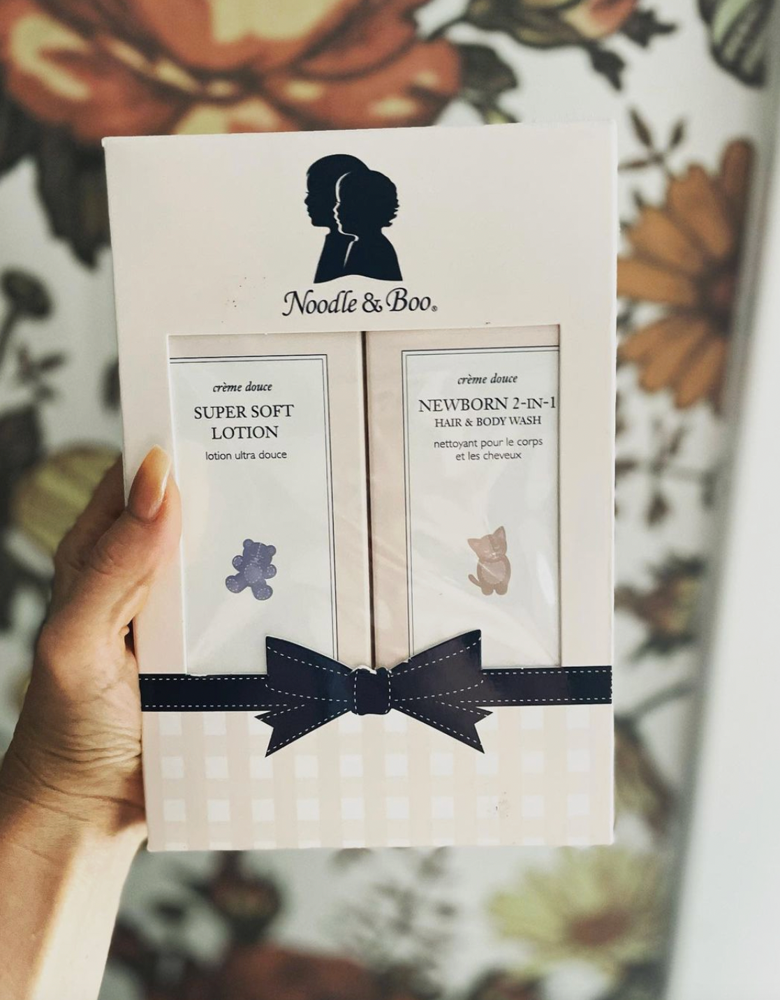 A hand holds a white Noodle & Boo gift set box featuring two tubes of baby care products: Super Soft Lotion and Newborn 2-in-1 Face & Body Wash, tied with a navy ribbon, against a blurred floral background.