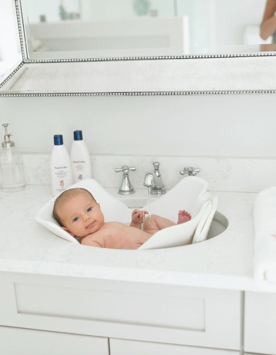 A baby lying on a white infant bath seat in a white bathroom sink vanity, with baby wash bottles and a mirror visible in the background.