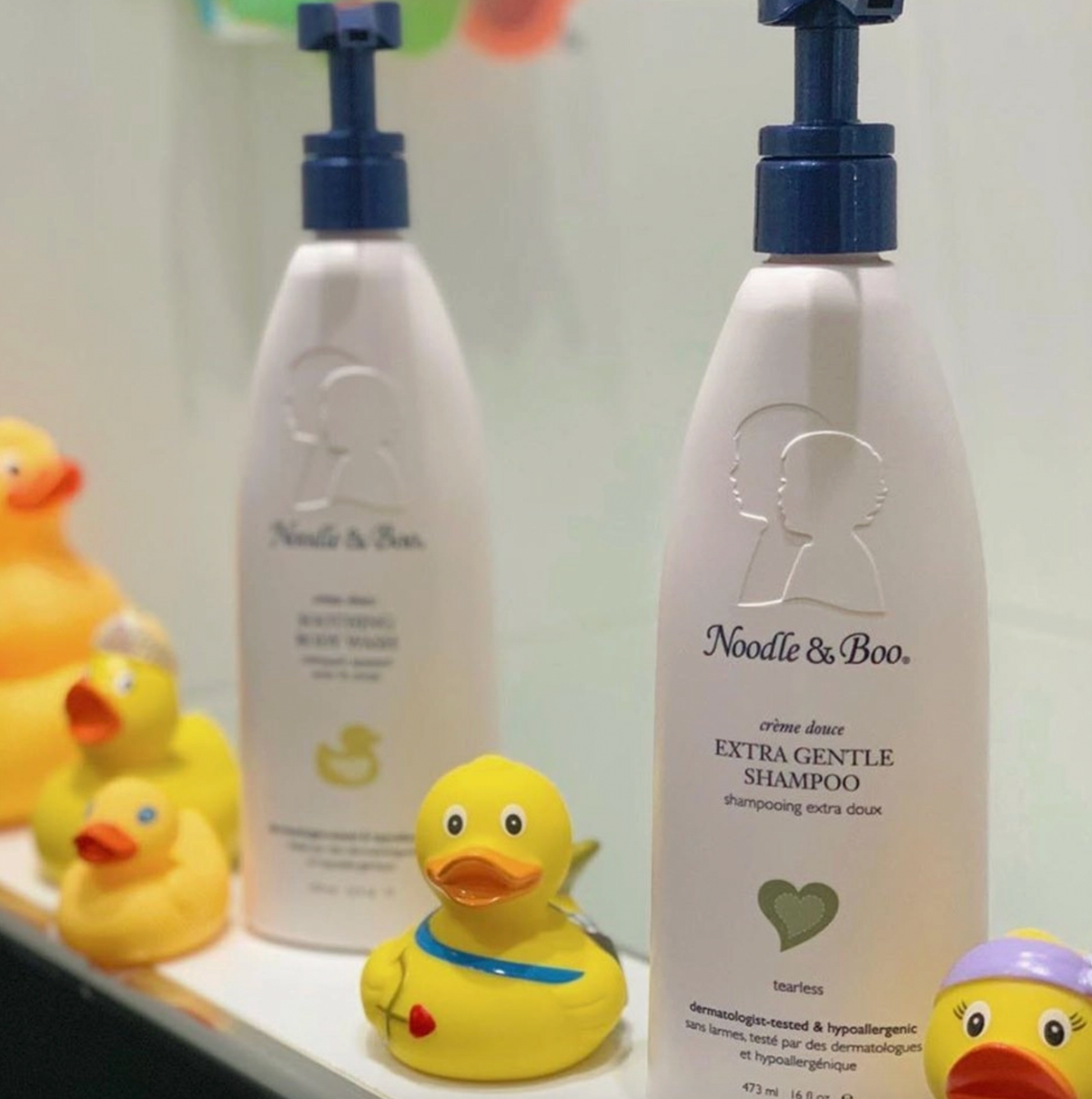 Two white pump bottles of Noodle & Boo baby shampoo and body wash displayed on a shelf with small yellow rubber ducks.