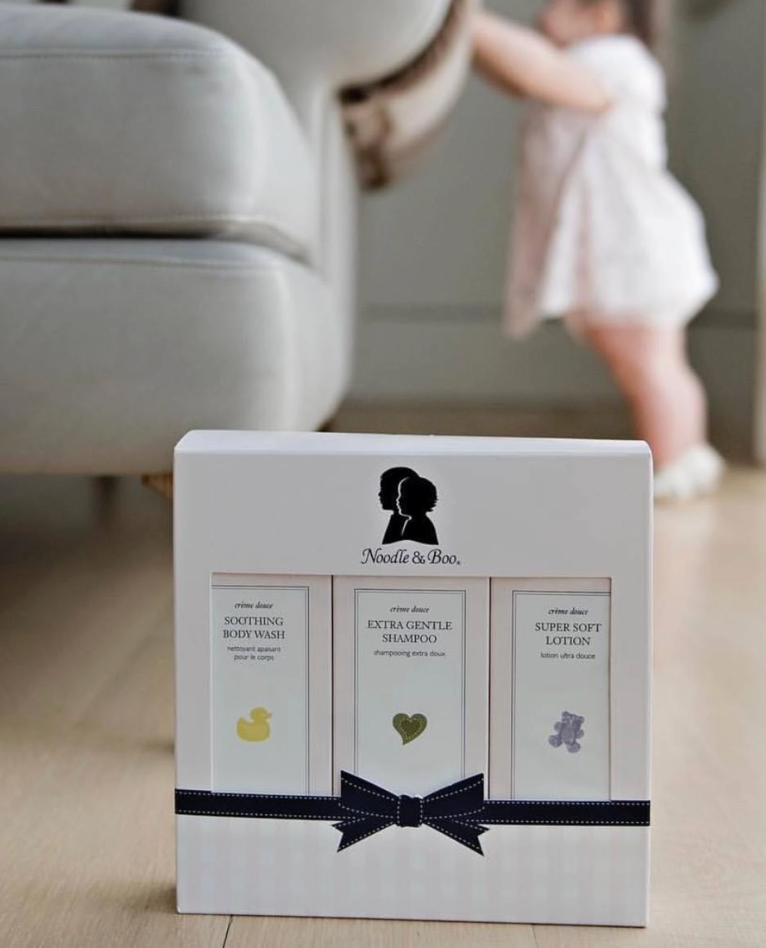 A boxed baby care set from Noodle & Boo sits on a light wood floor in a softly blurred nursery setting, with a child in a white dress visible in the background. The box contains three products: soothing body wash, extra gentle shampoo, and super soft lotion, with small yellow, green, and lavender icons on a white package tied with a dark ribbon.