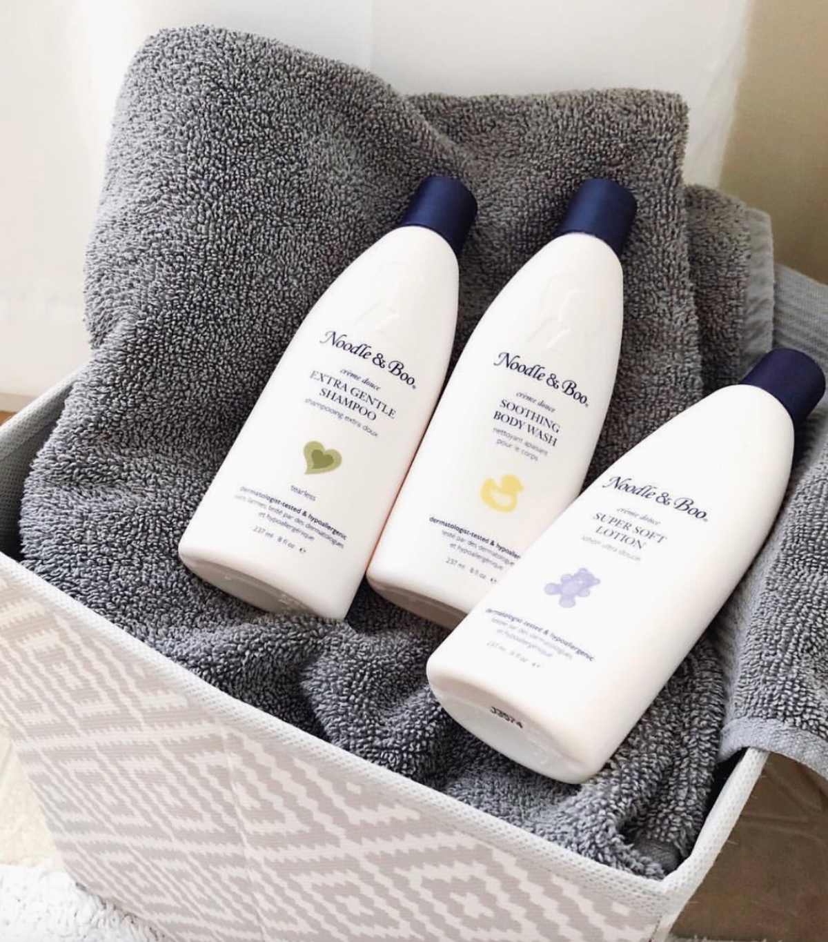 Three bottles of Noodle & Boo baby body wash arranged on a gray towel inside a white basket.