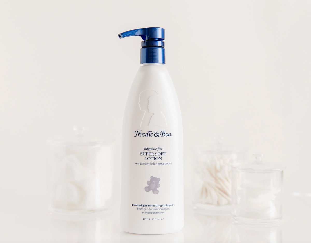 A white pump bottle of Noodle & Boo Super Soft Lotion with a blue pump, centered against a bright white background with softly blurred glass jars and cotton rolls.