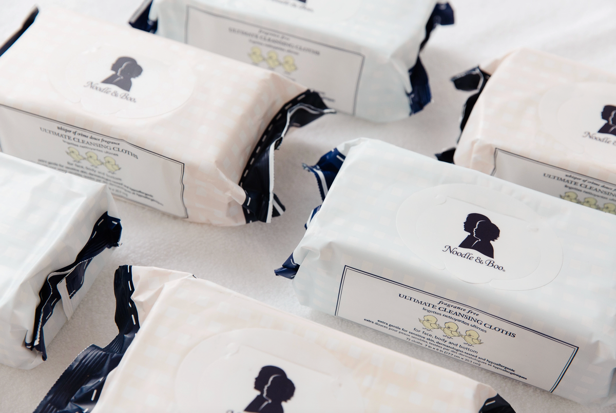 Several packs of cleansing cloths arranged on a white surface. The packs are off-white with dark navy blue ribbed edges and a black silhouette logo with the brand name.