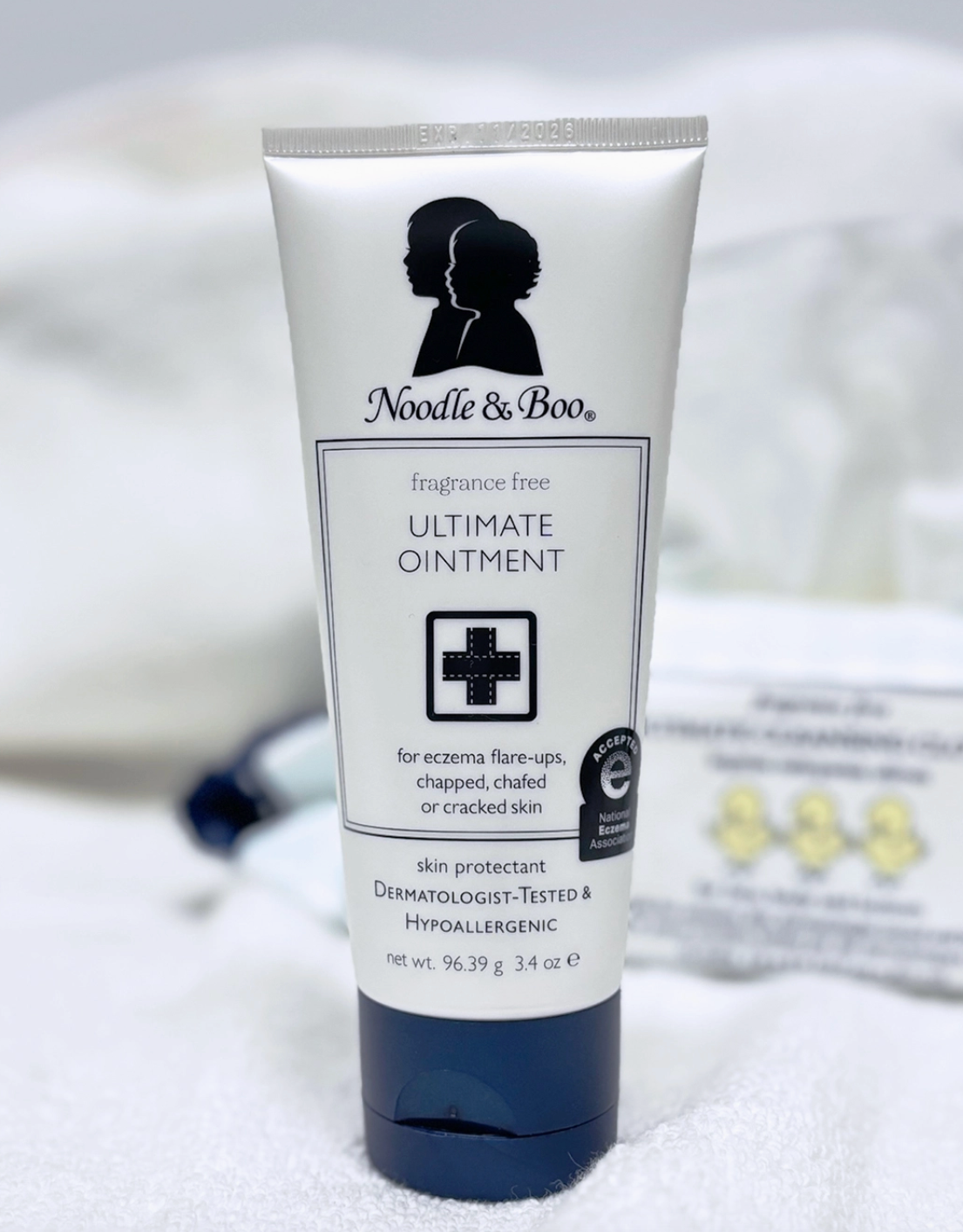 A white squeeze tube of Noodle & Boo Ultimate Ointment with a dark navy cap, standing upright on a white textured surface with a softly blurred background.