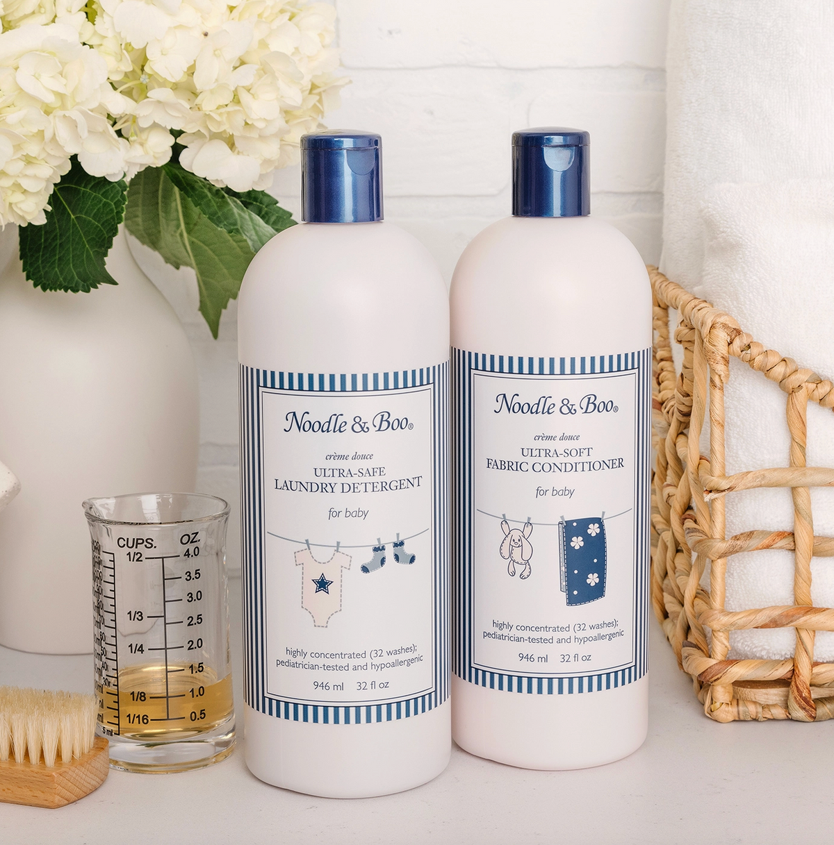 Two bottles of Needle & Boor baby laundry detergent and fabric conditioner standing on a white surface, styled with a glass measuring cup, white flowers, and a woven basket.