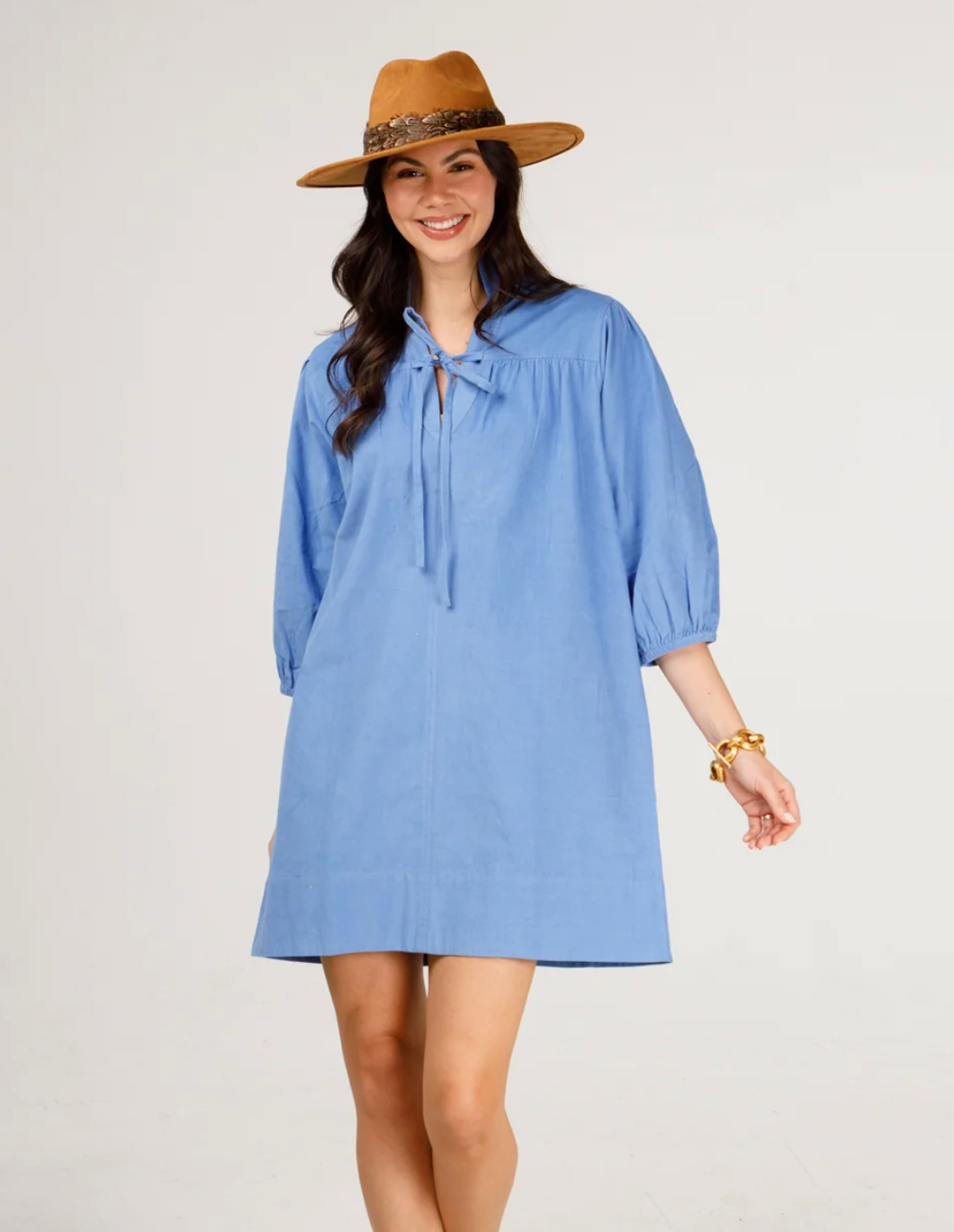 A woman modeling a heliotrope, long-sleeved mini dress with a tie-neck detail, styled with a brown wide-brim hat against a plain light gray background.