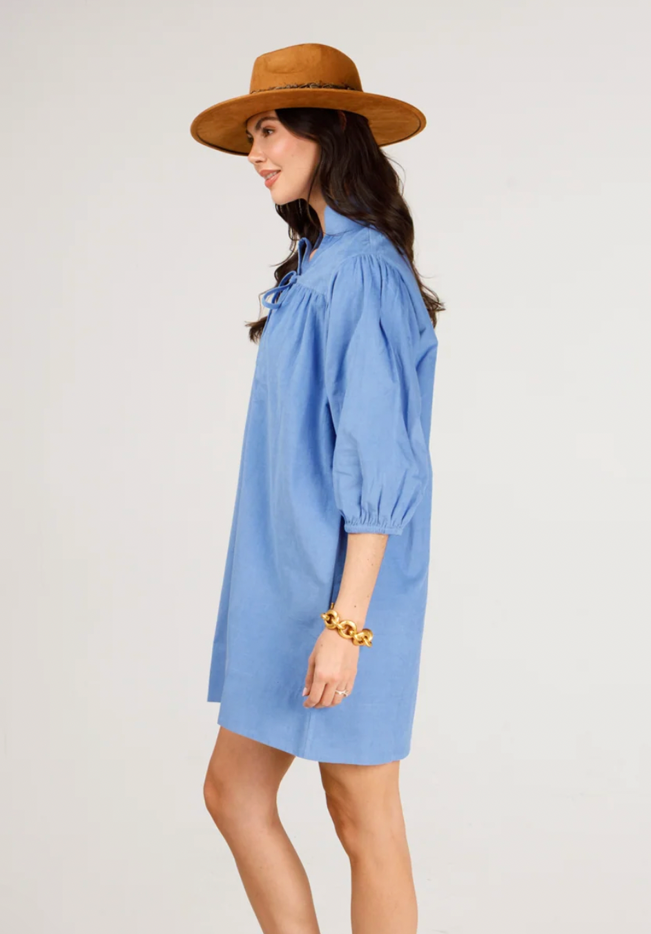 A woman wearing a heliotrope puff-sleeve shirt dress, a brown wide-brim hat, and a chunky gold bracelet, shown in profile against a light gray studio background.