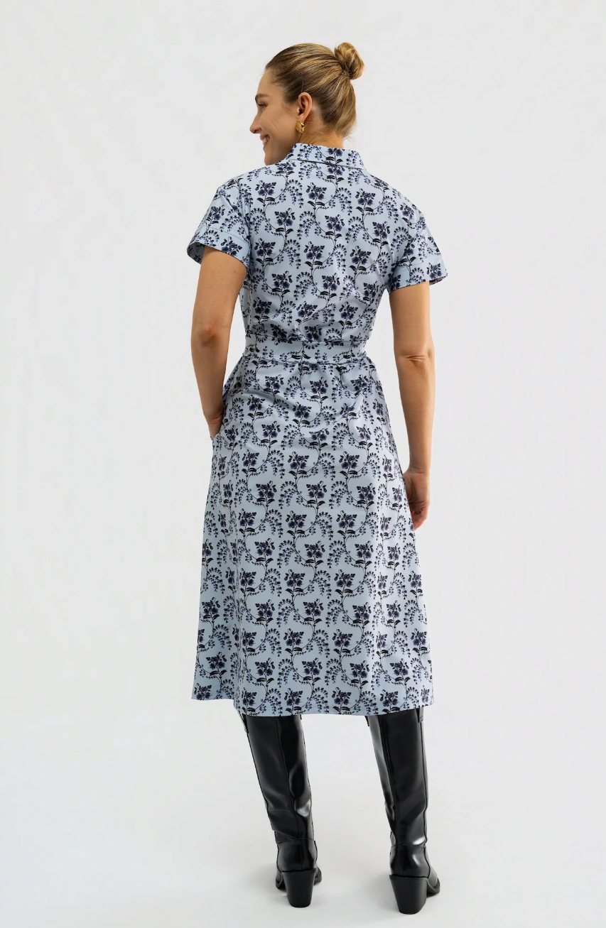 A woman is shown from the back wearing a short-sleeve midi dress with a light blue and navy floral block print, paired with black knee-high boots, against a plain light gray background.