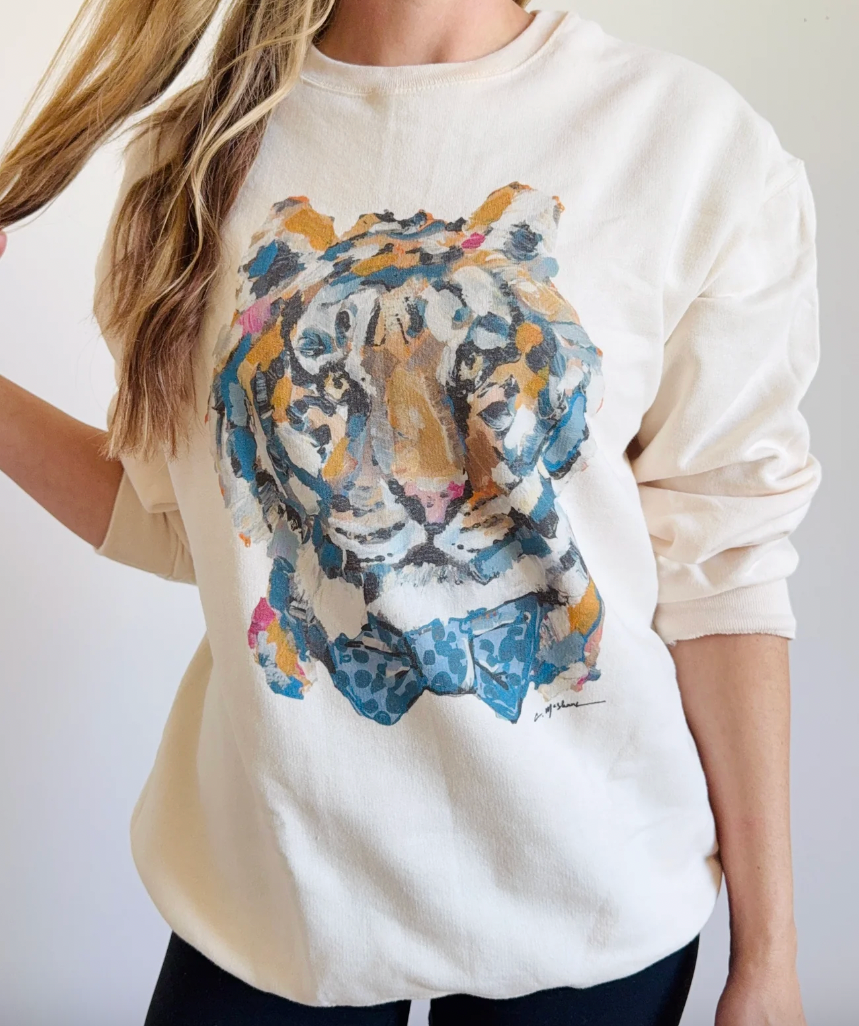 A woman wearing a cream-colored crewneck sweatshirt with a large, colorful acrylic tiger graphic on the front. The sweatshirt has long sleeves and is shown against a plain light background.