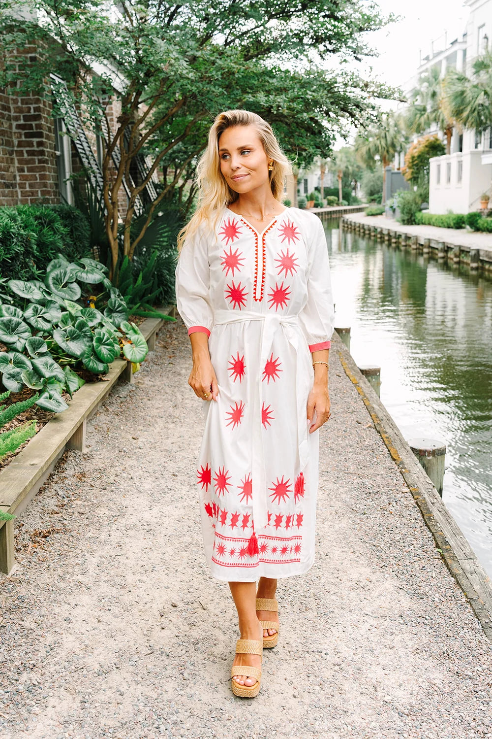 Woman in a white dress with red patterns walking along a path by a canal.